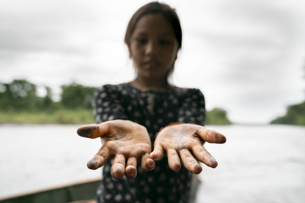 A girl holds out her hands dotted with oil from a spill contaminating the river behind her.