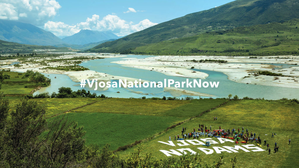 The Vjosa River in the background with surrounding mountains. In the foreground, a green field with a message that says "Vjosa No Dams".